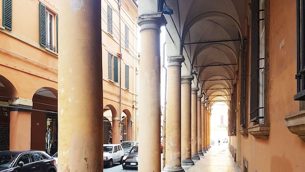 A corridor with columns and arches, lined with parked cars and buildings.