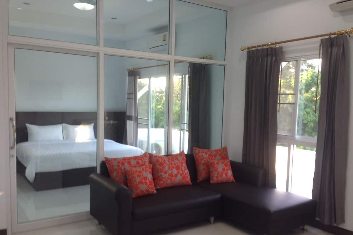A modern hotel room with a large bed, a black sofa with red pillows, and large windows offering a view of greenery.