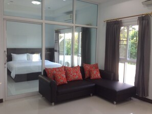 A modern hotel room with a large bed, a black sofa with red pillows, and large windows offering a view of greenery.