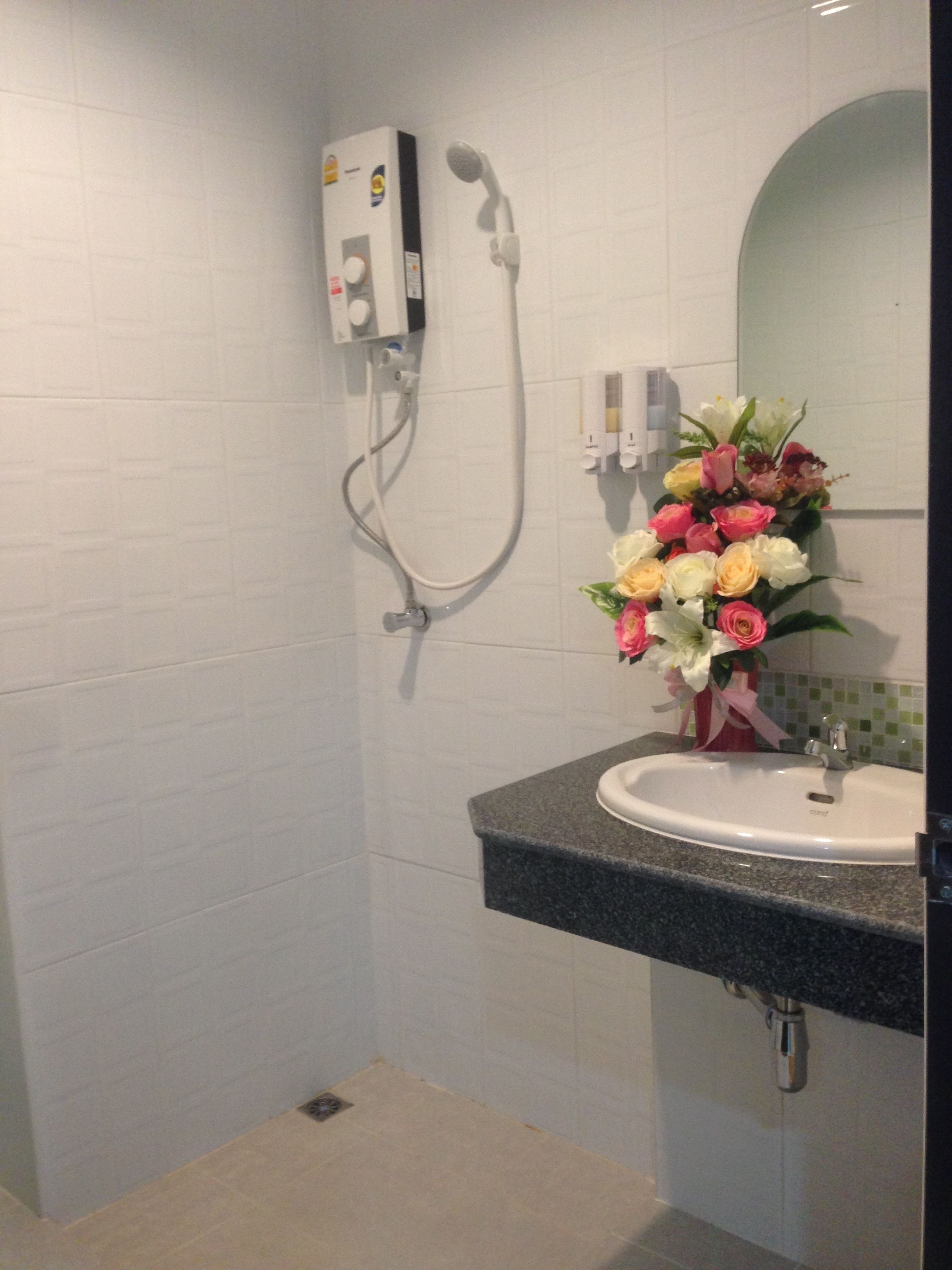 Une salle de bain avec un mur recouvert de carreaux blancs, une douche avec un pommeau de douche amovible, un lavabo avec un plan de travail en marbre et un vase de fleurs posé sur le plan de travail.
