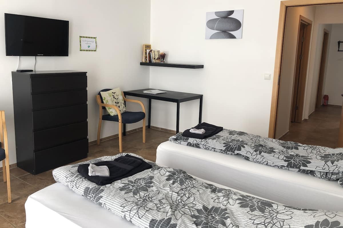 A room with two beds, a desk, a chair, a TV, and a shelf with books and a sign.