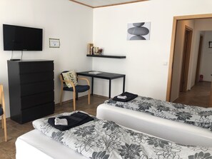 A room with two beds, a desk, a chair, a TV, and a shelf with books and a sign.