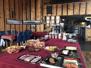 A buffet setup with various food items, including meats, breads, and fruits, in a rustic wooden room with tables and chairs. - Country Hotel Heydalur (Latur)