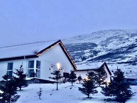 Una casa blanca con techo inclinado y esquina iluminada, rodeada de nieve en el suelo y árboles, con una montaña al fondo.