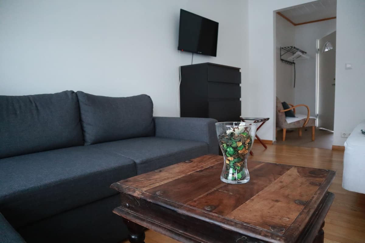 A living room with a dark grey sofa, a wooden coffee table with a glass vase, a black TV on the wall, and a white chair in the background.