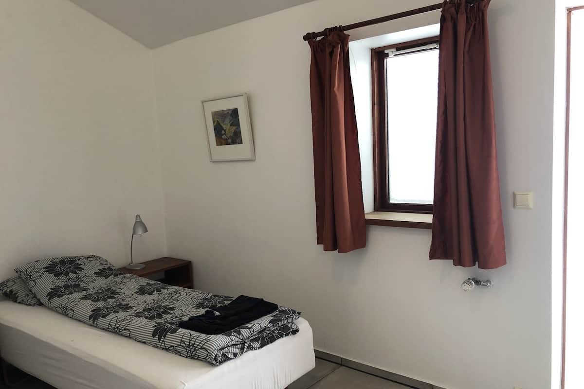 A small, simple room with a bed, a nightstand, a lamp, a window with curtains, and a wall-mounted picture.