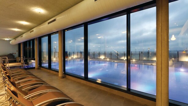 A swimming pool area with clear glass walls, a seating area with loungers, and a view of the pool through the glass.