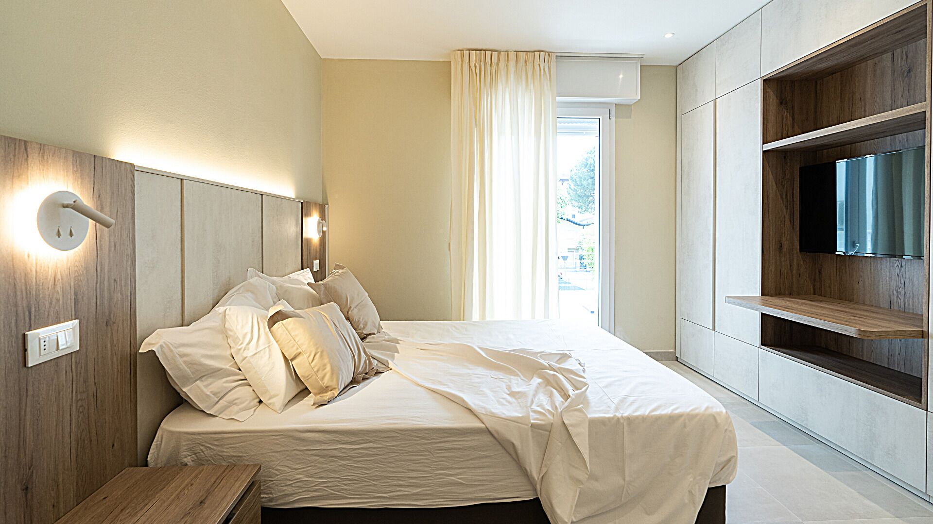 A modern bedroom with a large bed, built-in TV, and a window with curtains.