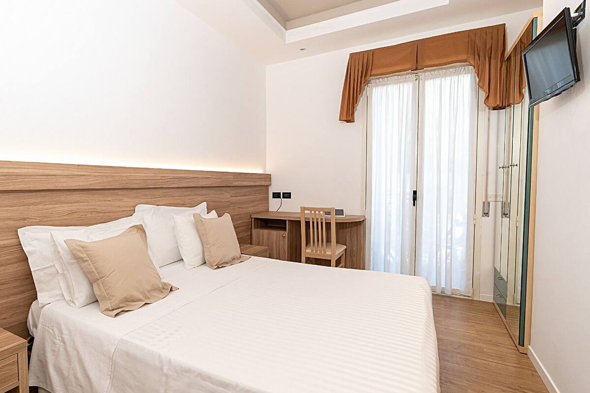 A hotel room with a wooden headboard, a bed with white linens, a bedside table with a phone, a desk with a chair, a TV mounted on the wall, and a sliding door with curtains.