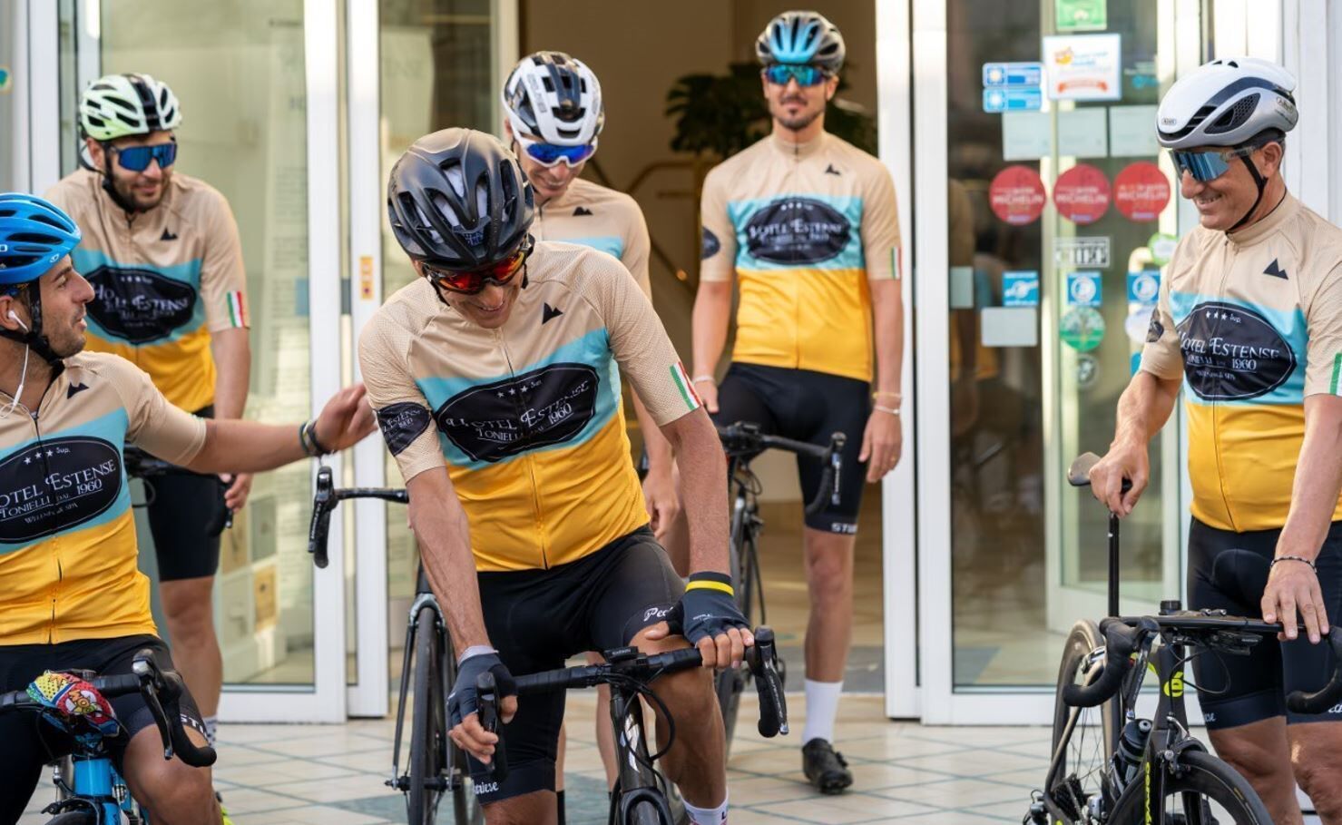En grupp cyklister i likadana uniformer står utanför en byggnad med glasdörrar och ett klinkergolv.