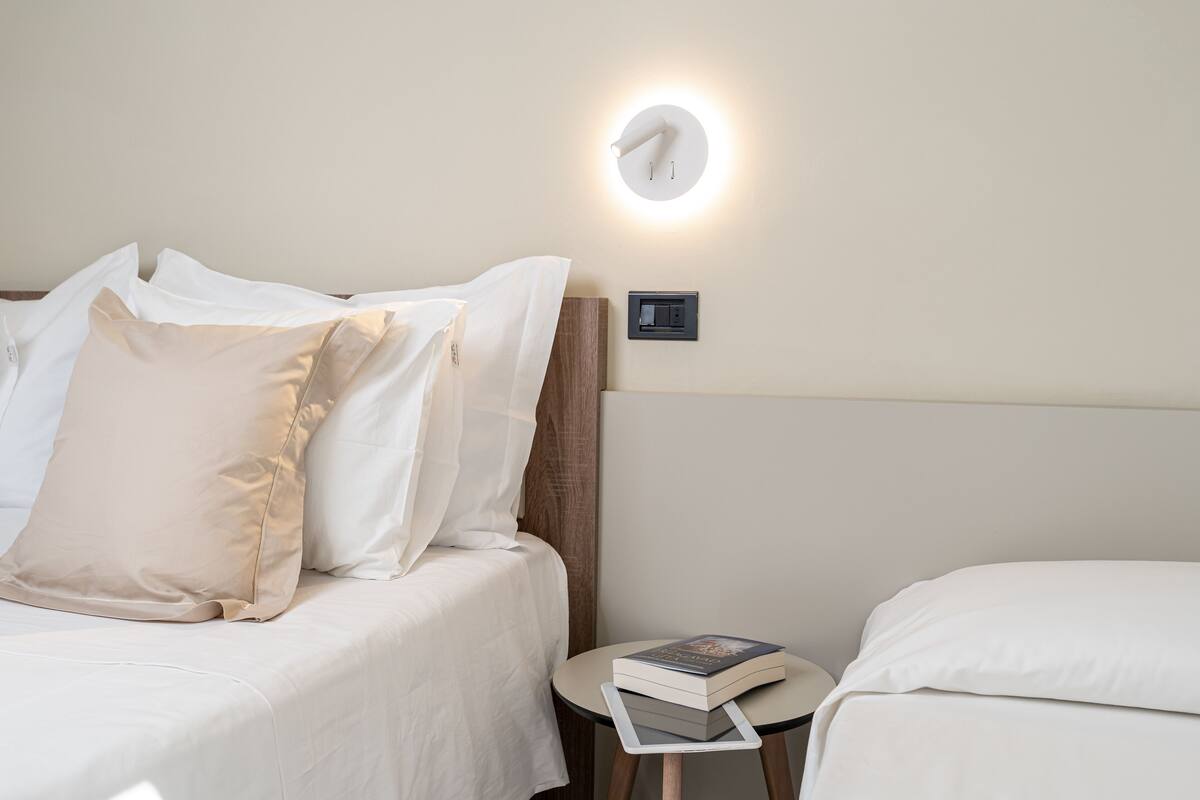 A neatly made bed with white bedding and a beige pillow, a bedside table with a book and a remote control, and a wall-mounted light fixture.