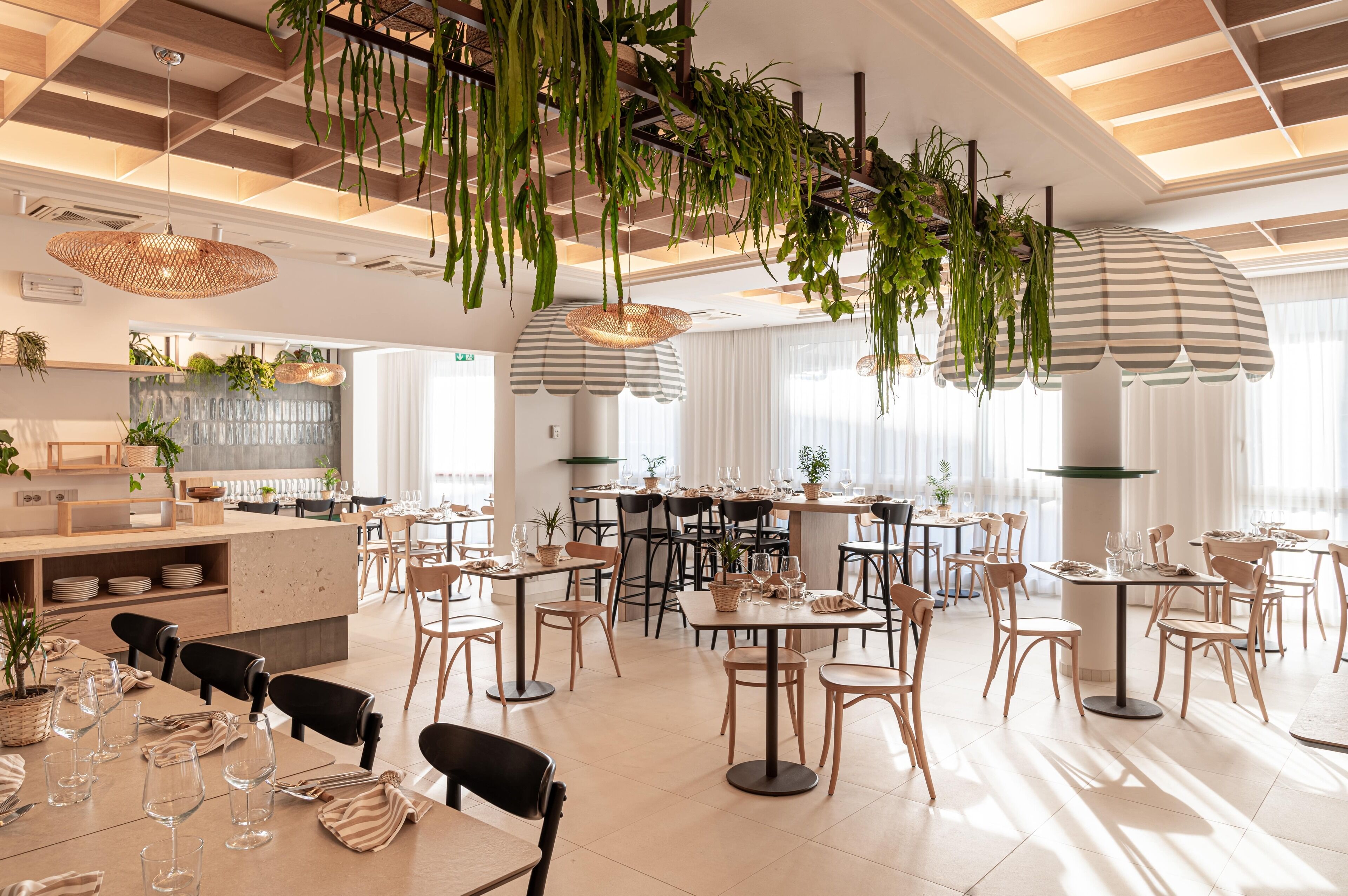 Un restaurante moderno con mesas y sillas de madera, plantas colgantes y sombrillas a rayas.