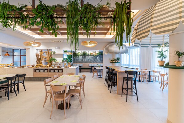 A modern restaurant with wooden tables and chairs, hanging plants, and a striped umbrella.