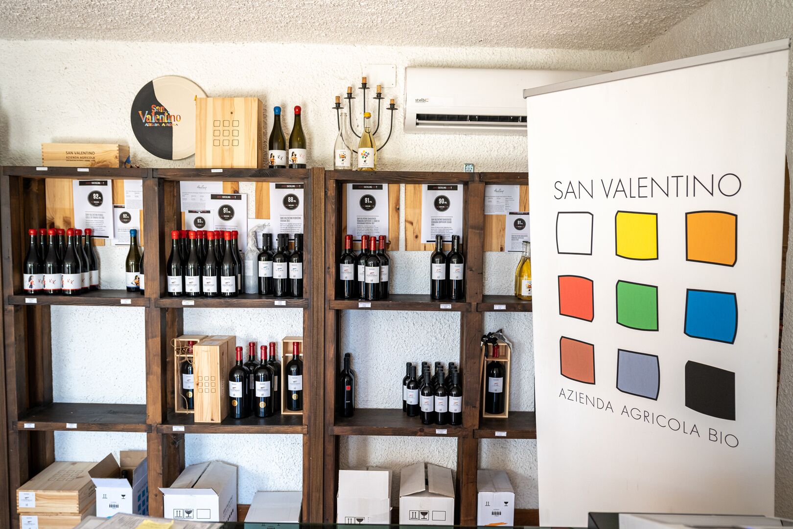 A wine shop with wooden shelves displaying various bottles of wine, a wooden sign with 'San Valentino' branding, and a whiteboard with color squares.