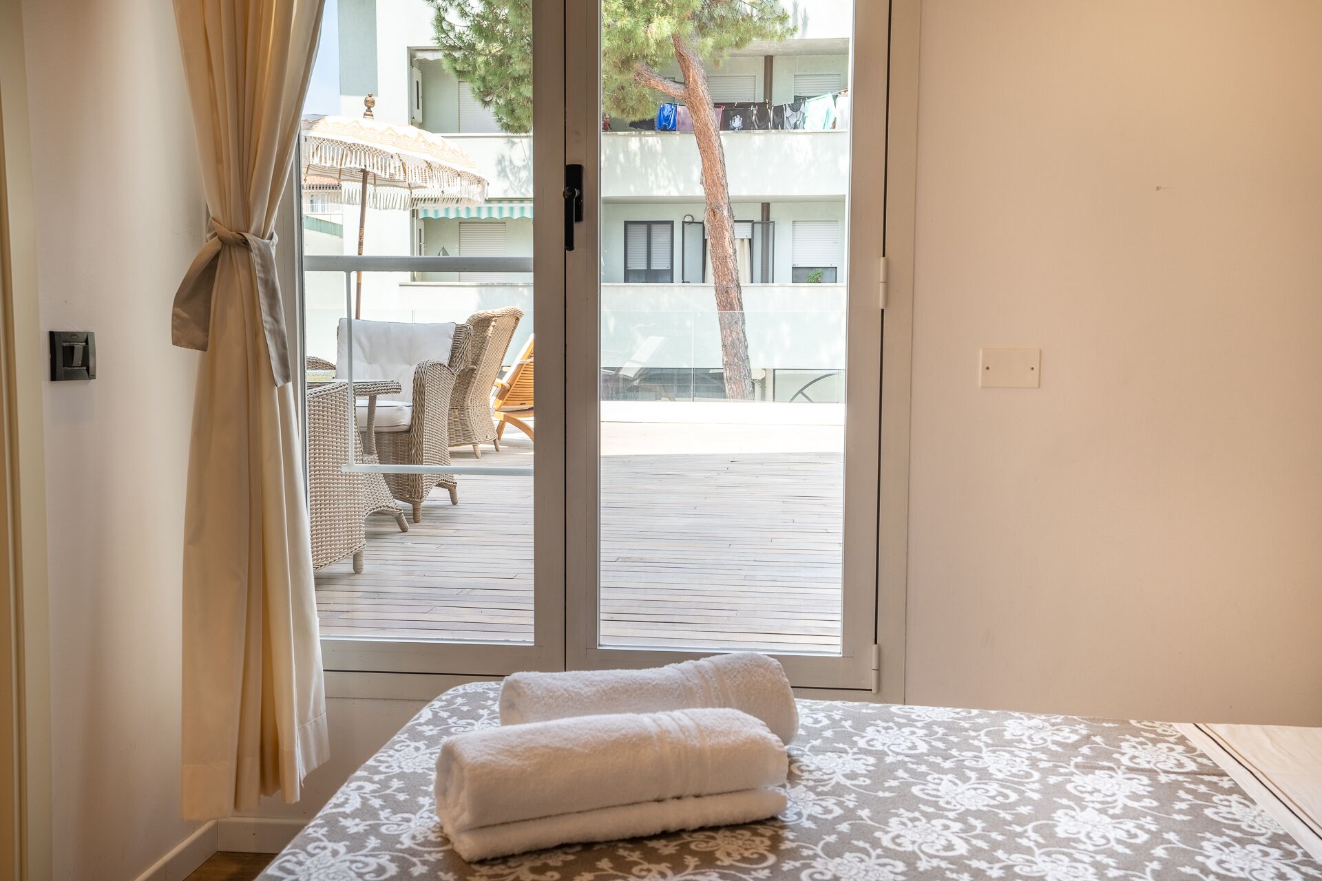 A hotel room with a bed, a window with curtains, and a view of an outdoor area with chairs and a tree.
