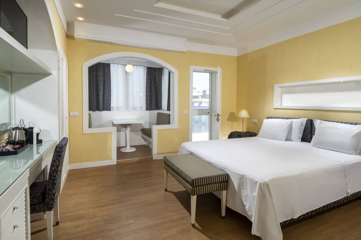A hotel room with a bed, a desk, a chair, a bench, a window with curtains, and a door.