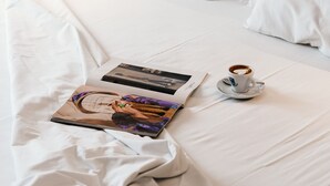 Una cama bien tendida con sábanas blancas, una revista y una taza de café sobre un platito.