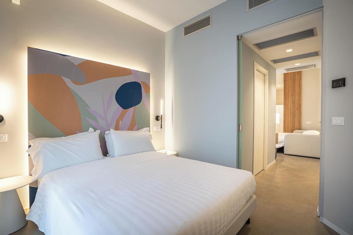 A hotel room with a large bed, a modern abstract painting on the wall, and a separate sitting area with a sofa and a wooden panel wall.