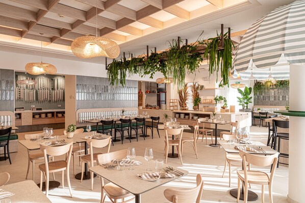 A spacious dining area with multiple tables and chairs, hanging plants, and a modern ceiling design. - Hotel Estense (Gatteo)