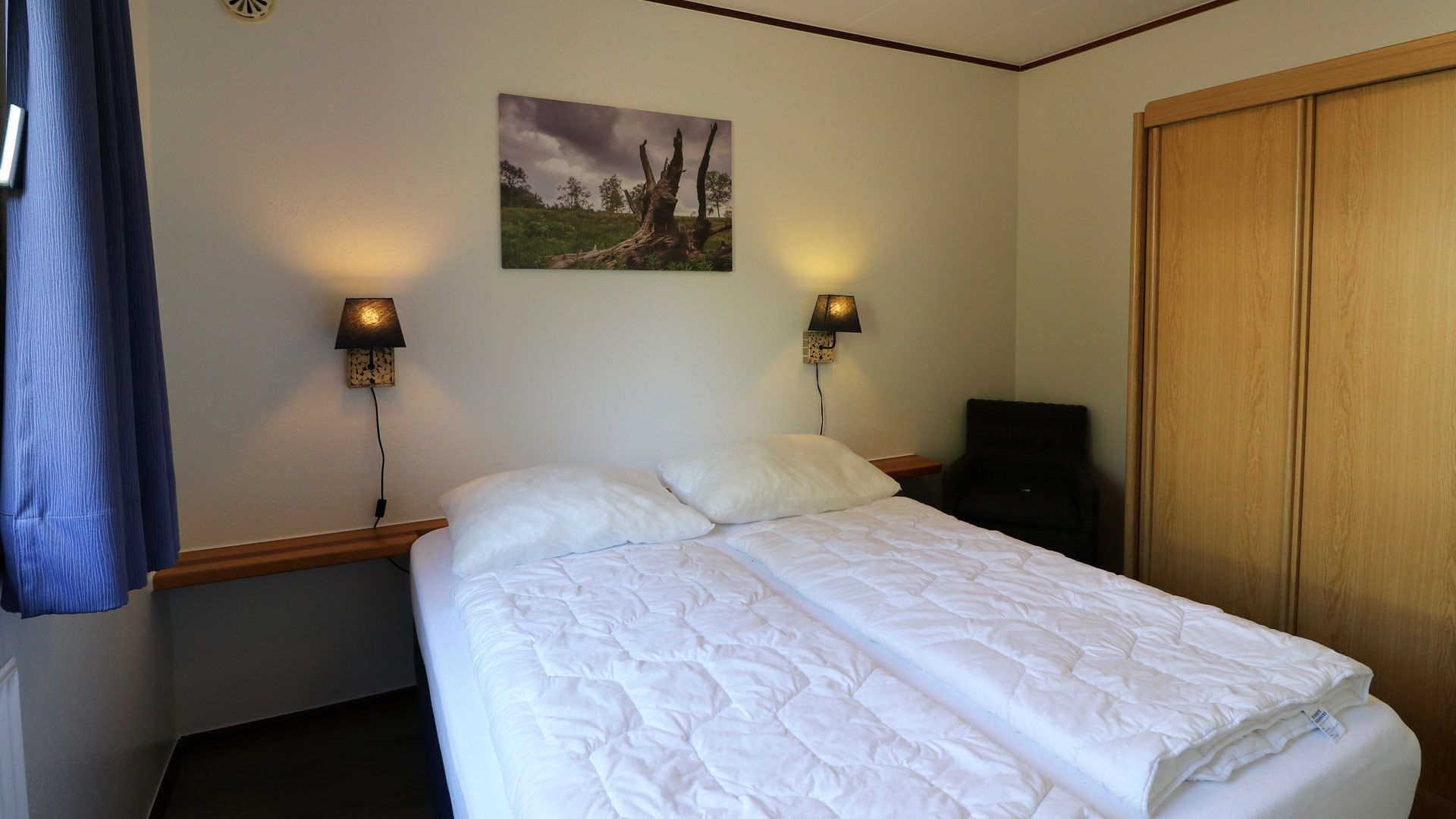 A hotel room with a bed, two wall-mounted lamps, a painting, a wardrobe, and a chair.