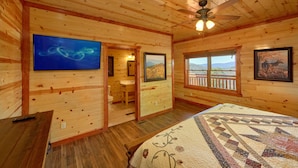 A wooden cabin bedroom with a bed, a ceiling fan, a TV, a bathroom, and a window with a mountain view.