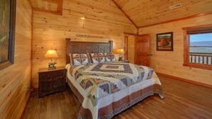 A cozy log cabin bedroom with a large bed, two bedside lamps, a wooden headboard, a framed picture, and a window with a view.