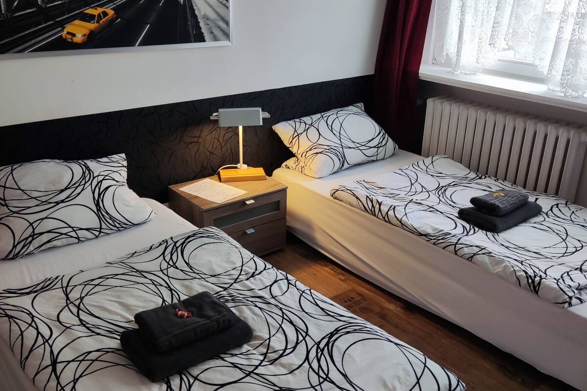Une chambre d’hôtel avec deux lits, une table de chevet, une lampe, un radiateur et un tableau représentant un paysage urbain accroché au mur.