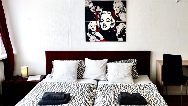 Een tweepersoonsbed met beddengoed met een patroon, een nachtkastje met een lamp en een muur met een pop-art schilderij.