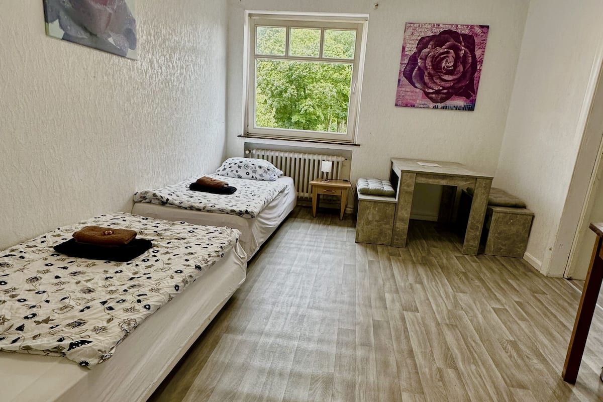 Une petite pièce propre et bien rangée, avec deux lits simples, un radiateur, une petite table et une fenêtre donnant sur un paysage verdoyant.