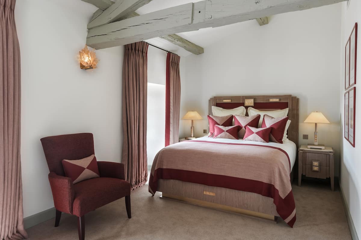 Ein Schlafzimmer mit einem großen Bett, zwei Nachttischen mit Lampen, einem Sessel und einem Fenster mit Vorhängen.