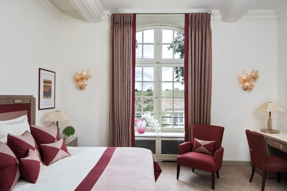 Ein Hotelzimmer mit einem Bett, zwei Stühlen, einem Schreibtisch, einem Fenster mit Vorhängen, einem gerahmten Bild an der Wand und einer kleinen Pflanze auf dem Nachttisch.