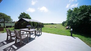 An outdoor patio with a table and chairs, a wooden structure in the background, and a grassy field with sheep.