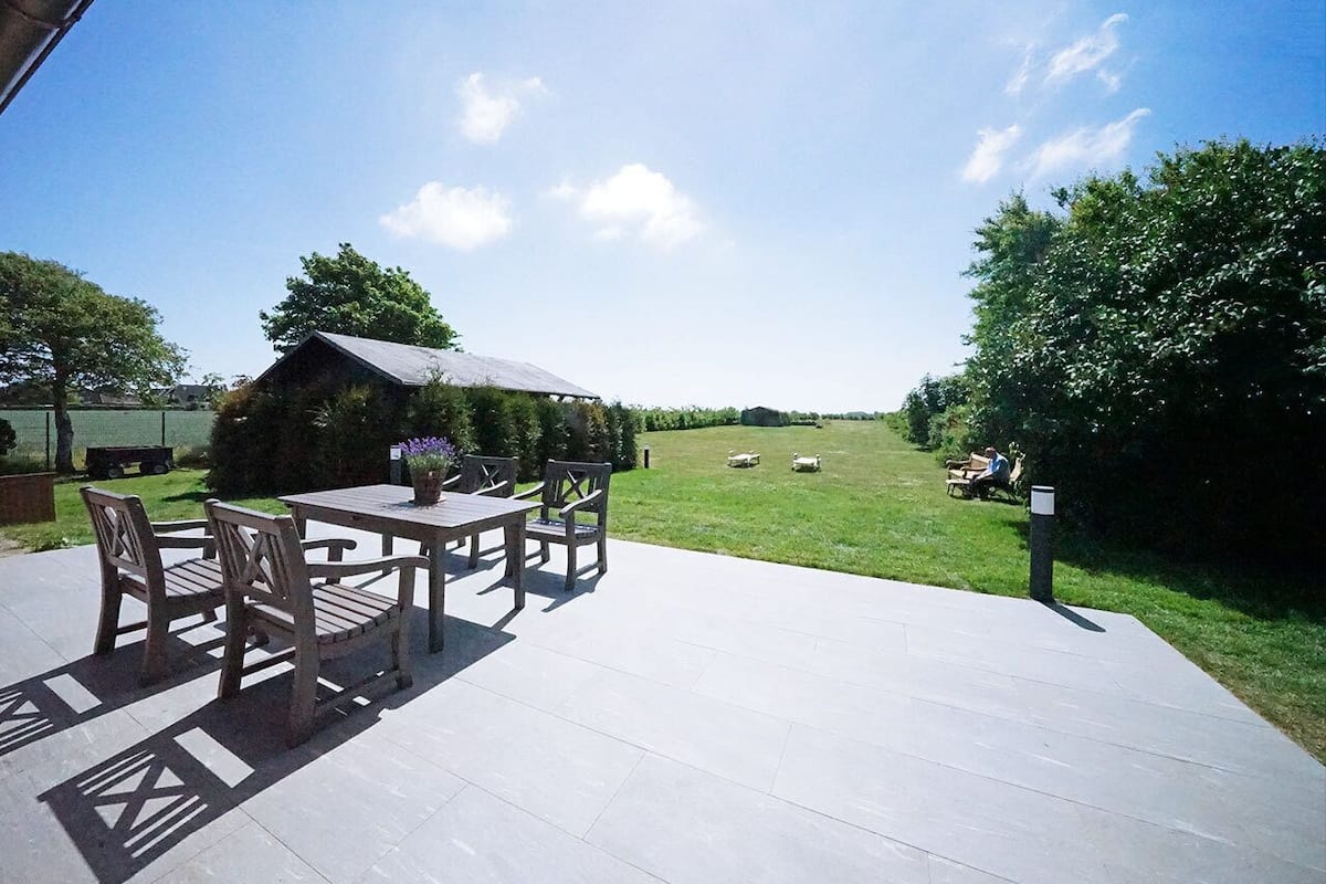 Une terrasse extérieure avec une table et des chaises, une structure en bois en arrière-plan et un champ verdoyant où paissent des moutons.
