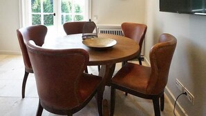 A dining area with a round table and six brown leather chairs. There is a window with a view of greenery outside, a wall-mounted clock, and a flat-screen TV on the wall.