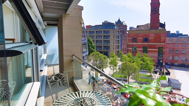 A balcony with a glass railing overlooking a cityscape, featuring historic buildings and a clear sky. - Live in Leeds Millenium Square Apartment (Leeds)