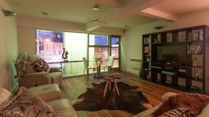 A modern living room with a large window, a fireplace, and a bookshelf. - Live in Leeds Millenium Square Apartment (Leeds)