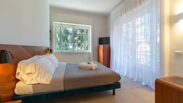 A neatly made bed with white linens, a wooden headboard, and a green upholstered footboard. There is a television on a stand to the right of the bed, and a window with white curtains to the left.
