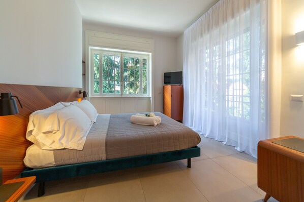 A neatly made bed with white linens, a wooden headboard, and a green upholstered footboard. There is a television on a stand to the right of the bed, and a window with white curtains to the left. - Le Stanze del Lago Suites & Pool (Como)