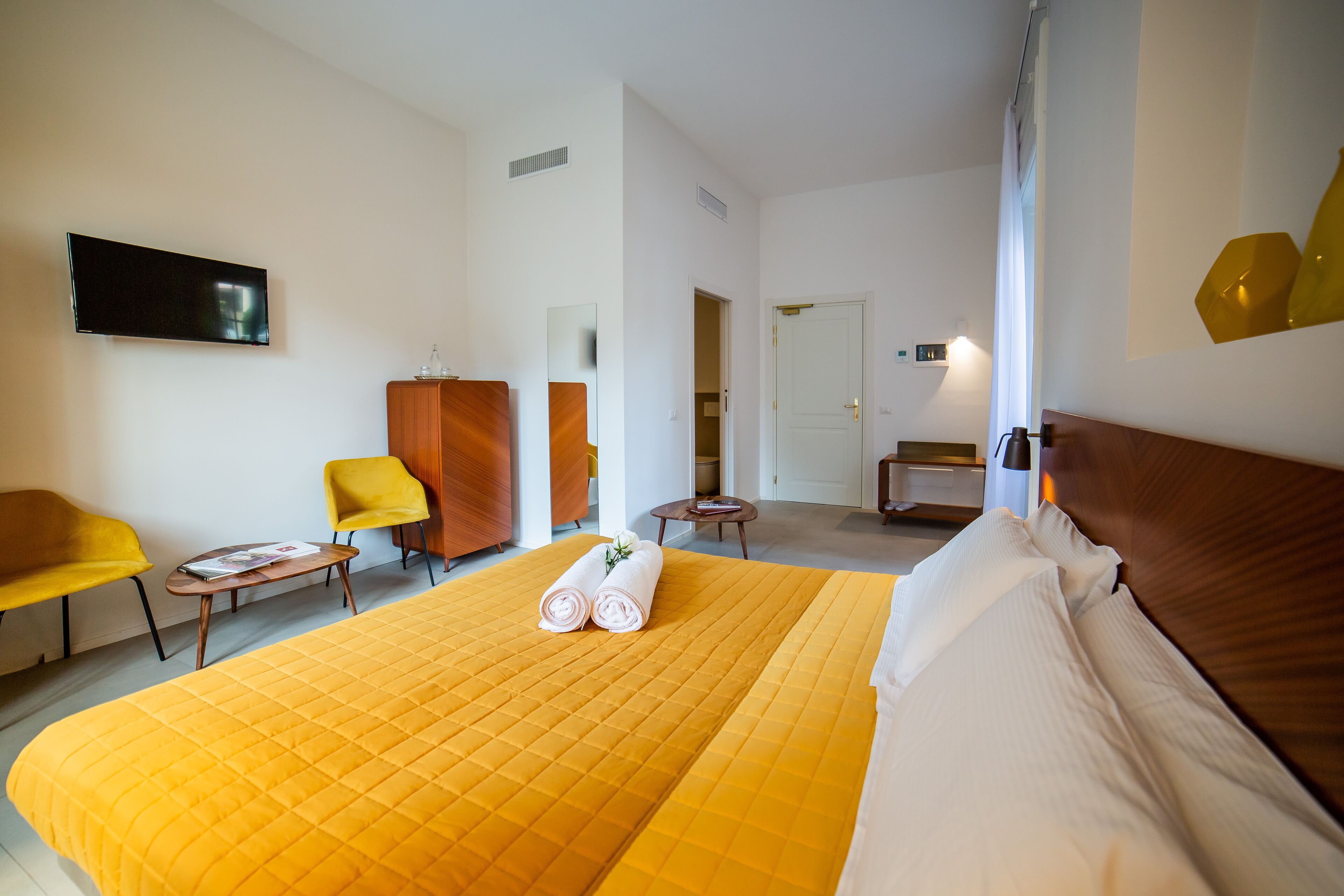 A hotel room with a large bed, a TV mounted on the wall, a wooden wardrobe, a small table with a magazine, and two chairs.