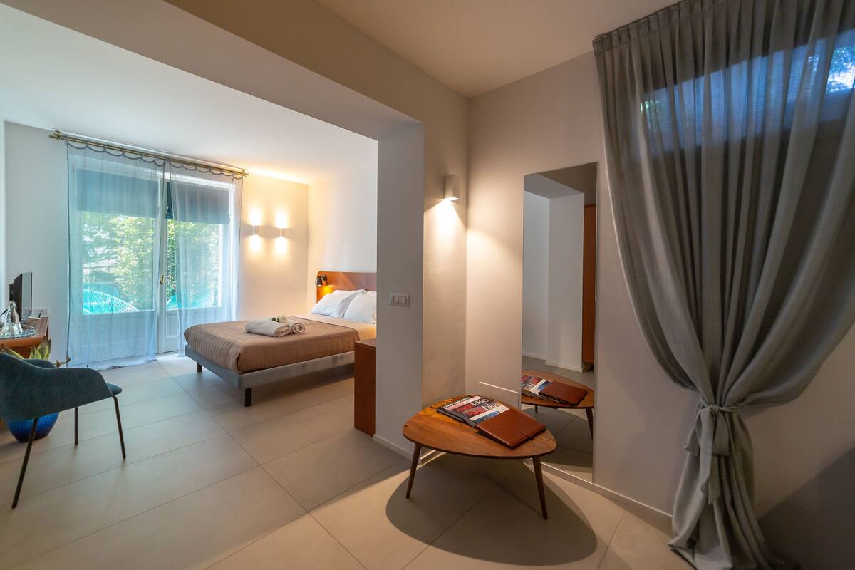 Una habitación de hotel moderna con cama, mesita de noche, silla y un ventanal grande con cortinas.