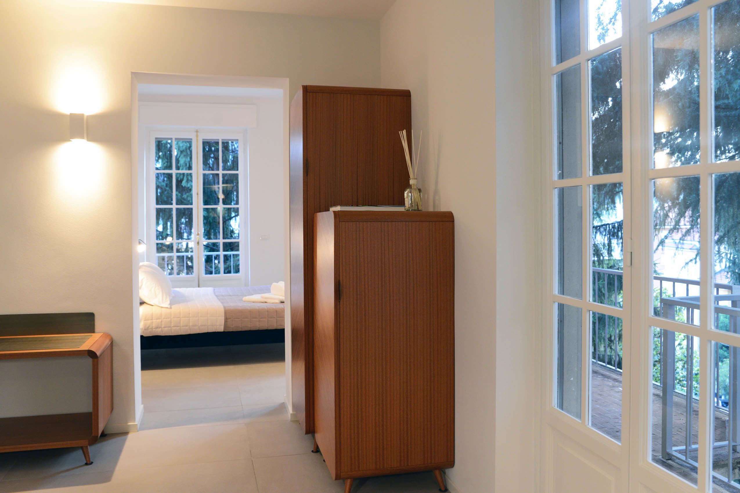 A bedroom with a bed, a wooden wardrobe, a nightstand, and a window with a view of trees.