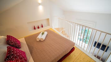 Camera da letto con un letto, cuscini, un comodino, una piccola mensola con oggetti decorativi, una finestra e una ringhiera da balcone.