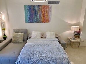 A modern bedroom with a grey sofa, a patterned bedspread, and a colorful abstract painting on the wall. - Casares Beach Golf Apartment With Private Garden and Pool Access (Casares)