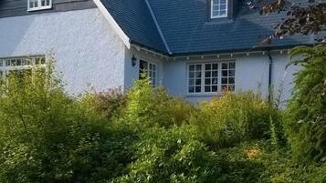 A two-story house with a sloped roof, surrounded by a well-maintained lawn and dense shrubbery.