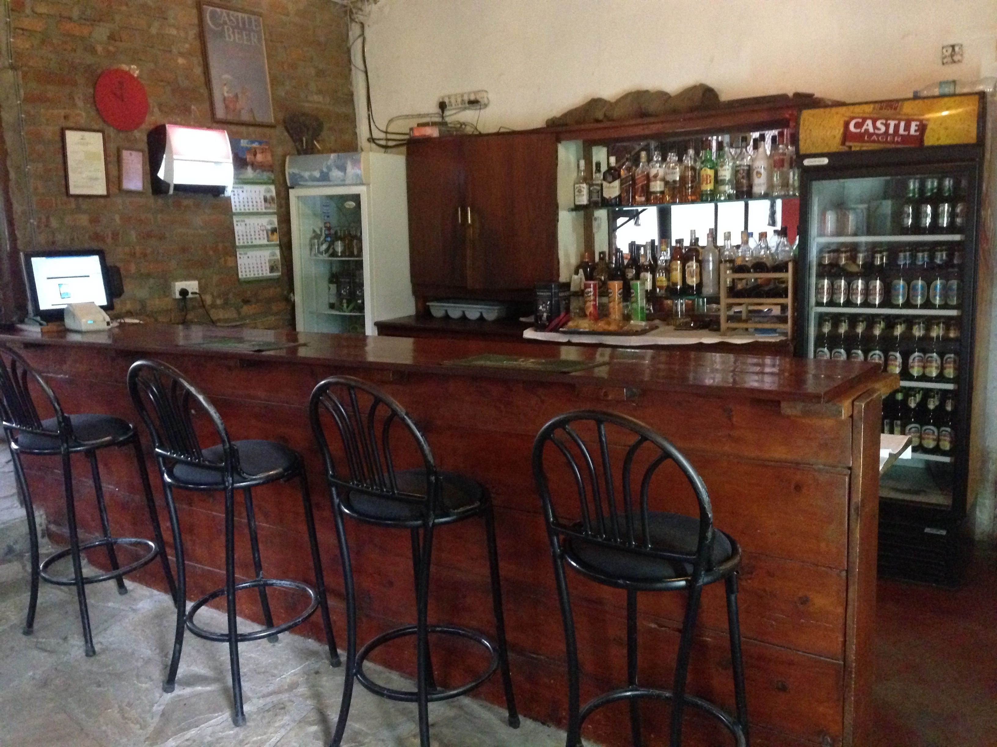 A bar area with a counter, bar stools, a Castle Lager refrigerator, and various bottles and glasses.
