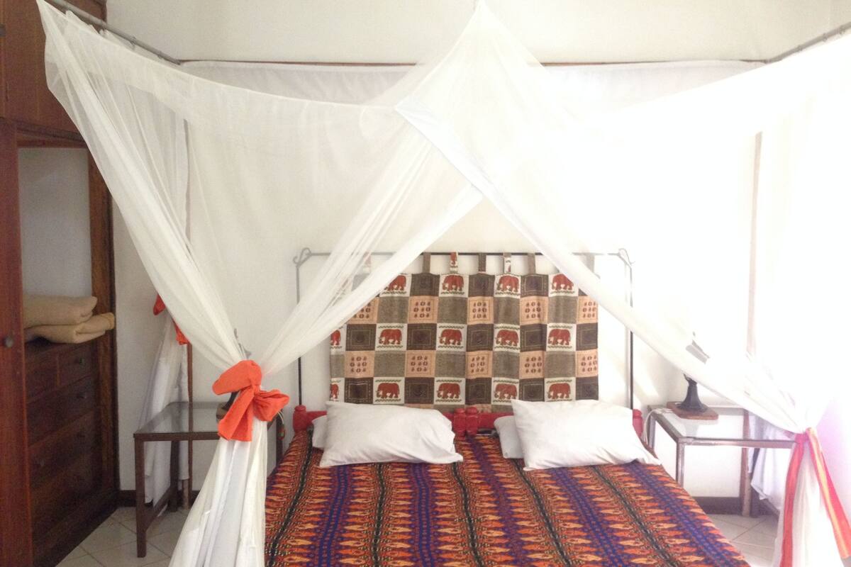 Une chambre avec un lit placé sous un baldaquin blanc, une tête de lit décorée et une table de chevet avec une lampe.