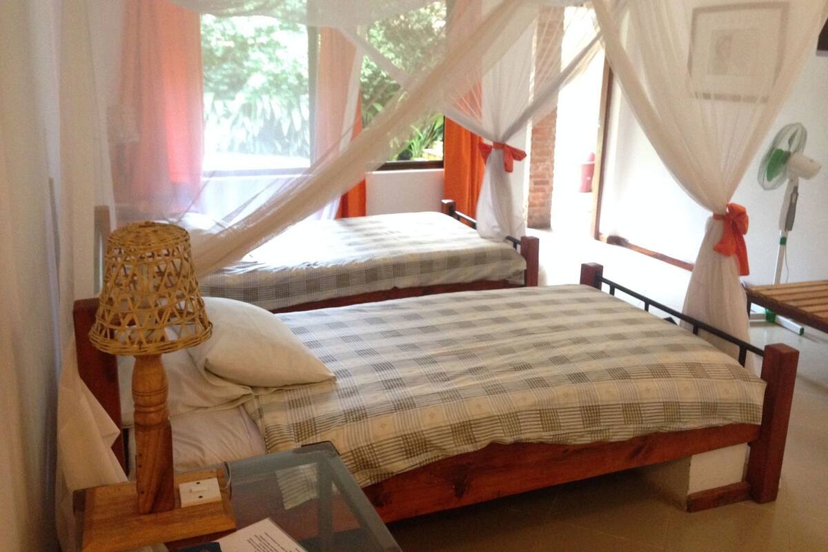 Une chambre avec deux lits, une table avec une lampe et une fenêtre avec des rideaux.