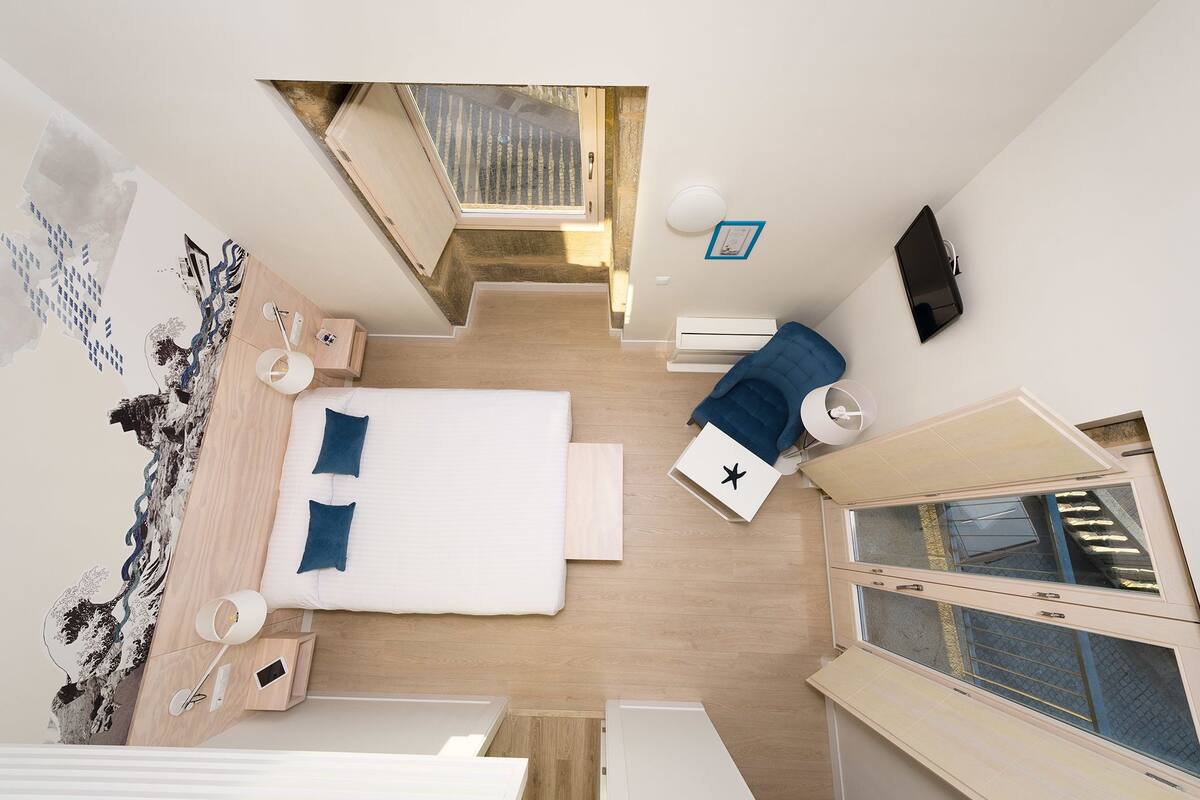 Ein kleines Hotelzimmer mit einem Bett, Nachttischen, einem Schreibtisch, einem Stuhl, einem Fernseher und einem Fenster.