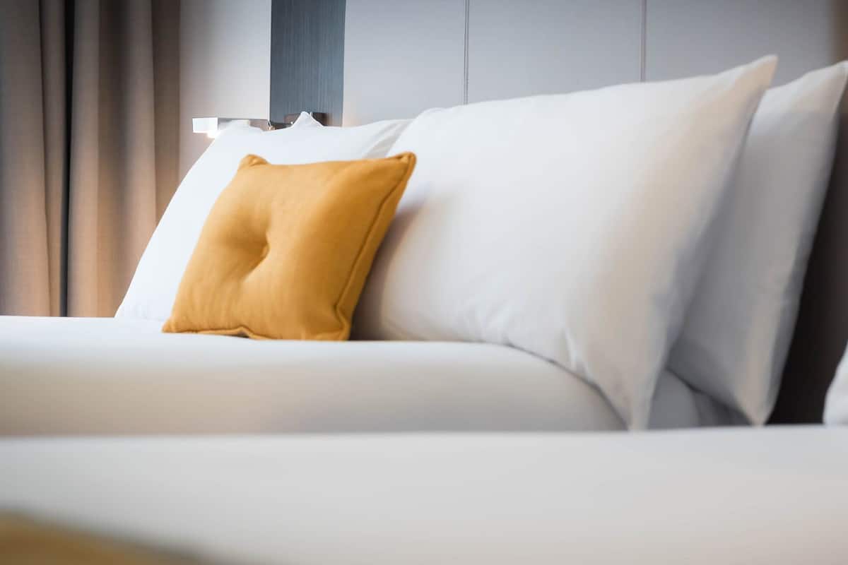 A modern hotel room with a neatly made bed, a single mustard-colored pillow, and a minimalist design.
