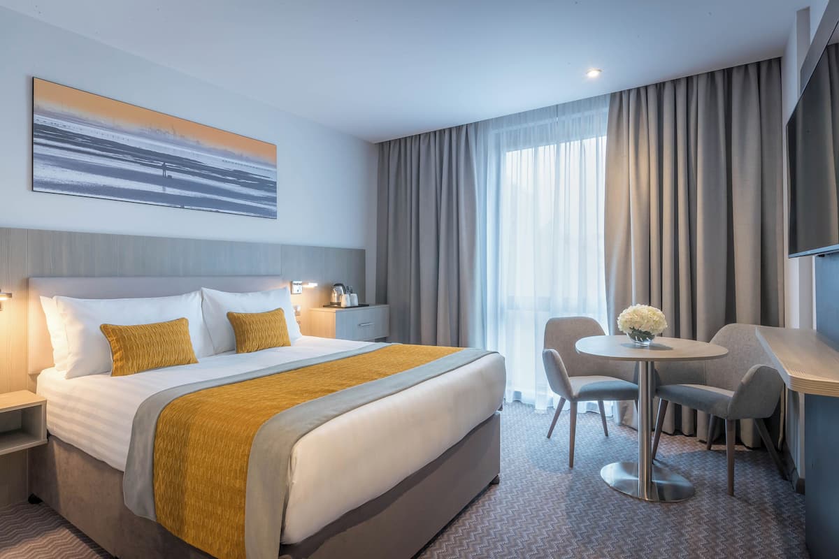 A hotel room with a large bed, a desk with two chairs, a small table with flowers, and a TV on the wall.